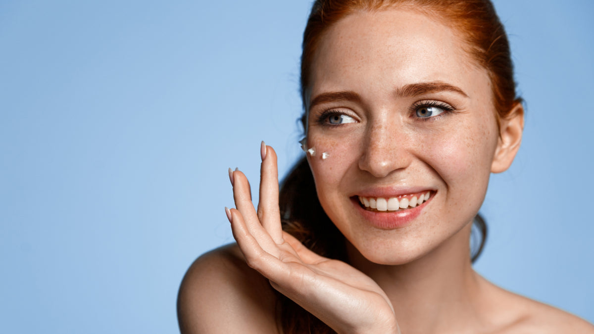 redhead girl on blue background with eye cream on glowing skin