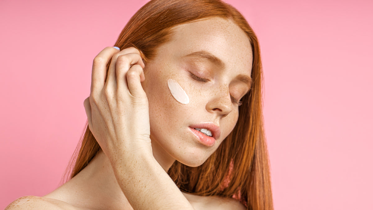 beautiful redhead touching skin, moisturizer on cheek, pink background