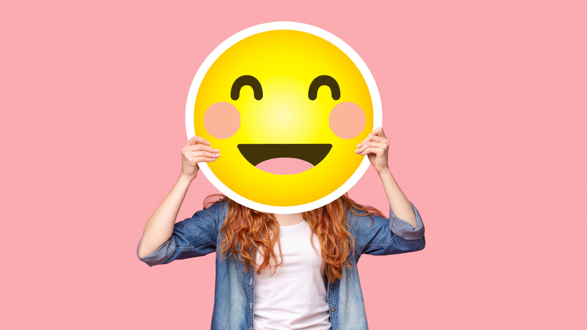 Redhead on pink background hiding large, smiling emoji in front of face