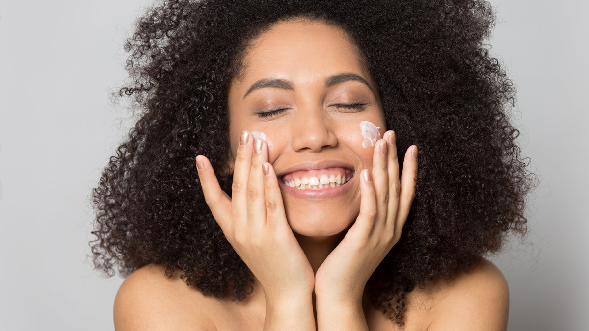 black girl applying moisturizer, smiling on gray background